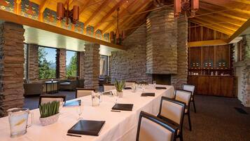 A dining area with stone walls, wooden beams, and a fireplace. Tables are set with white tablecloths, chairs, and glassware.