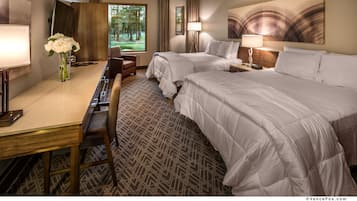 A hotel room with two beds, a desk, a chair, a lamp, and a window with a view of trees.