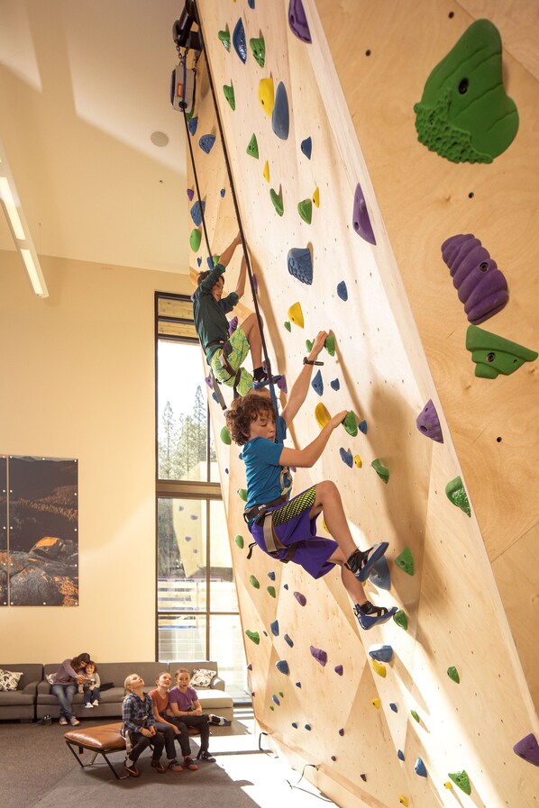 A climbing wall with colorful holds, a wooden climbing ladder, and a group of people observing from a couch. - The Inn at Nakoma (Clio)