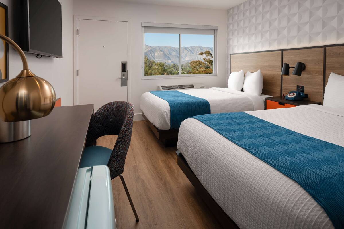A hotel room with two beds, a desk with a lamp, a chair, a window with a mountain view, and a wall with a geometric pattern.