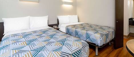 Habitación de hotel con dos camas, cada una con ropa de cama estampada, suelo de madera y una lámpara fijada a la pared.