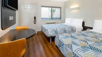 A hotel room with two beds, a table, a chair, a TV mounted on the wall, and a window with a view of greenery.