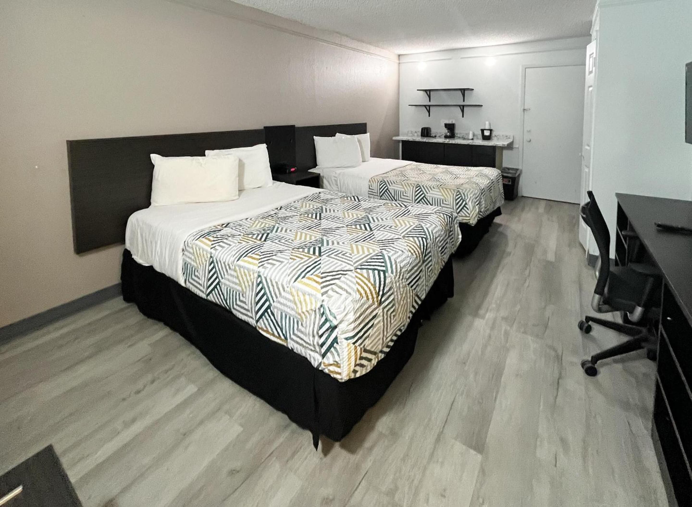 A hotel room with two beds, a desk, a chair, a small kitchenette, and a door.