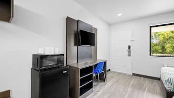 A compact hotel room with a flat-screen TV mounted on the wall, a small refrigerator, a desk with a chair, and a bed with a patterned bedspread.