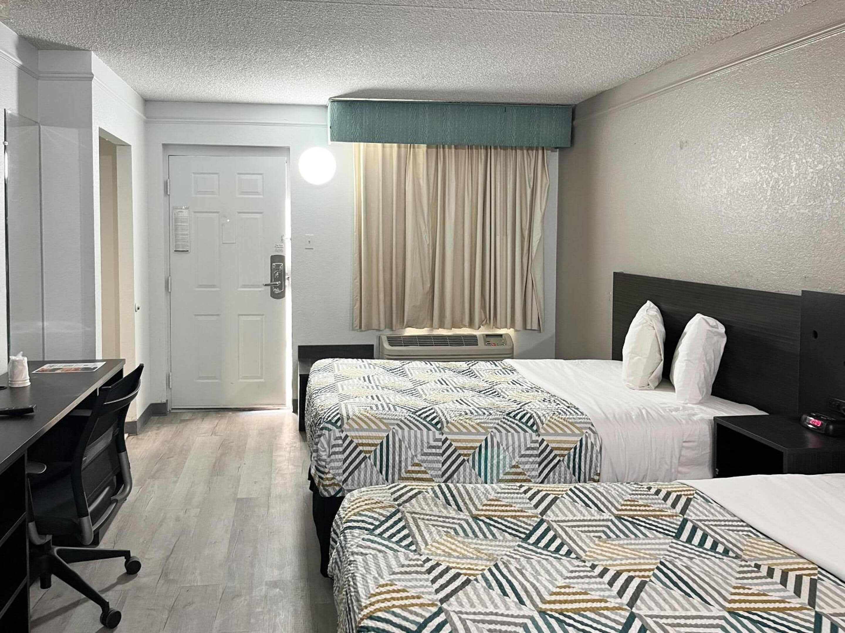 A hotel room with two beds, a desk, a chair, a door, and a window with curtains.