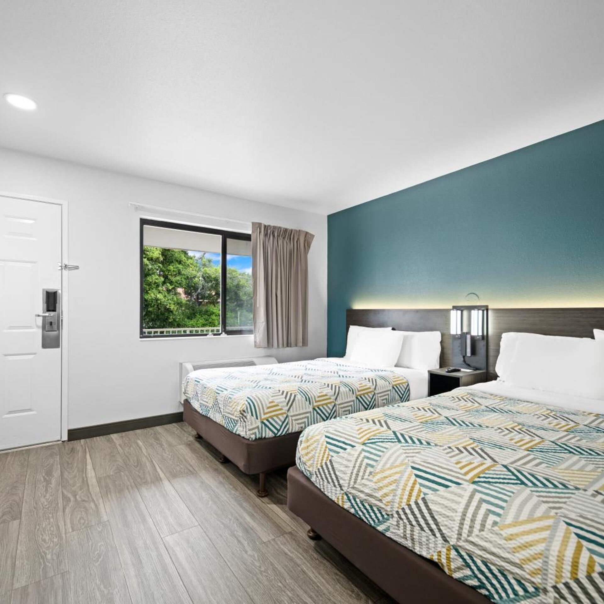 A hotel room with two beds, a window with a view of greenery, a door, and a nightstand with a lamp.