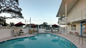A swimming pool with lounge chairs and umbrellas, surrounded by a white fence and a building with a balcony. - Vagabond Inn Monterey (Monterey)