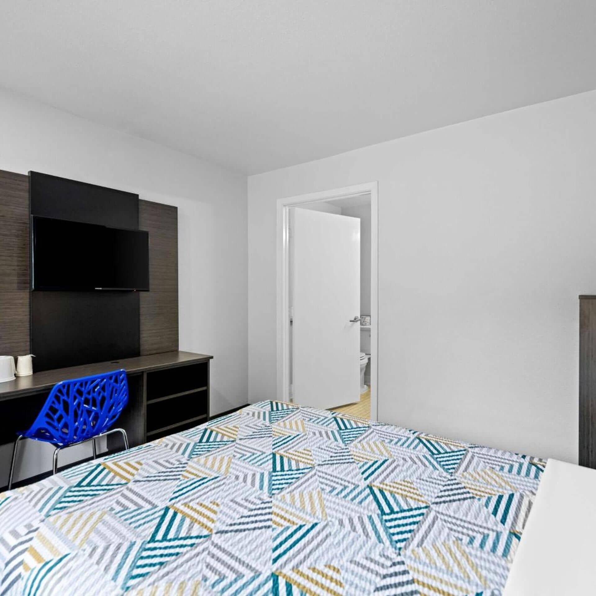 A bedroom with a bed, a desk, a chair, a TV, and a door leading to a bathroom.