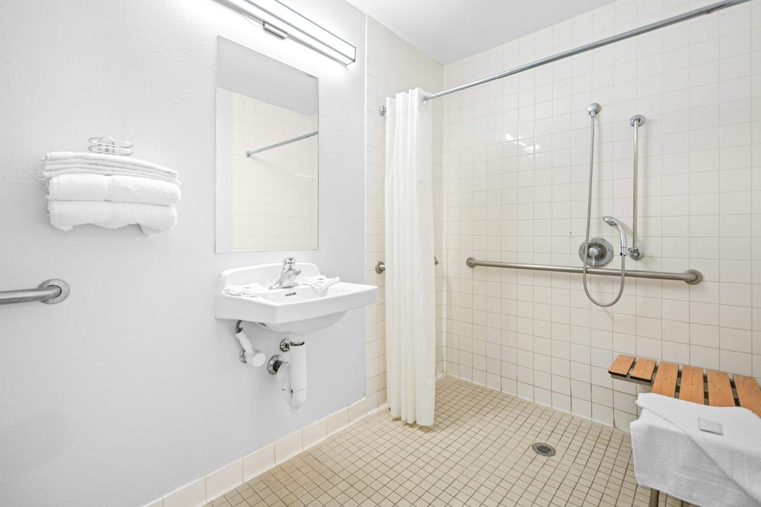 A bathroom with a sink, mirror, white towels, a shower with a grab bar, and a wooden bench.