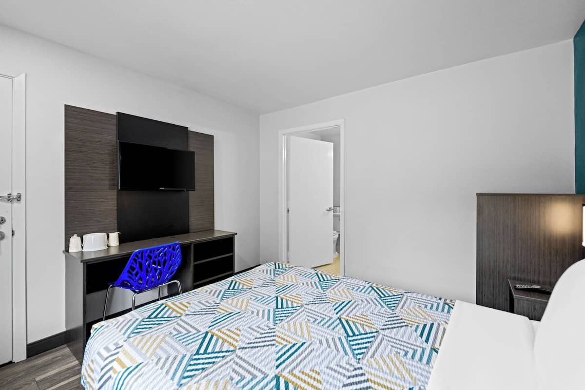A modern hotel room with a bed, a desk with a flat-screen TV, a blue chair, and a wardrobe.