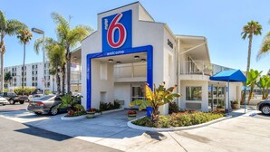 Un motel moderno con un llamativo letrero con el número '6', un acceso cubierto y un estacionamiento con coches y palmeras.