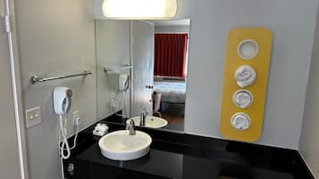 Une salle de bain d’hôtel avec un comptoir noir, un lavabo blanc, un miroir, un porte-serviettes avec des serviettes et un rideau rouge en arrière-plan.