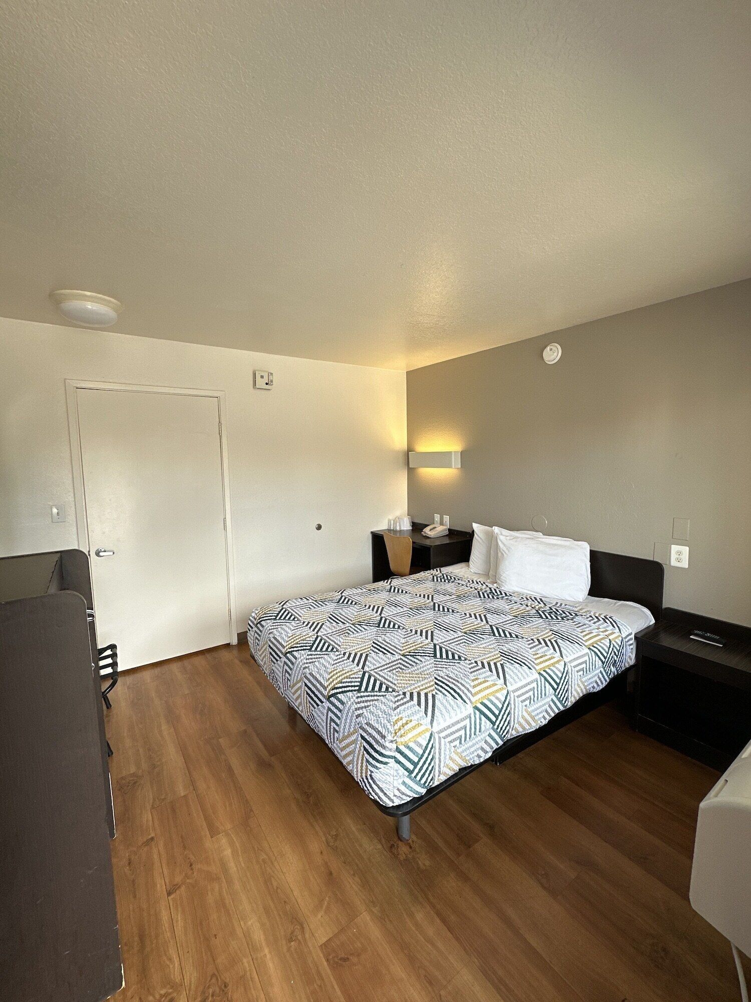 A hotel room with a bed, a desk, a chair, a nightstand, and a wall-mounted light fixture.
