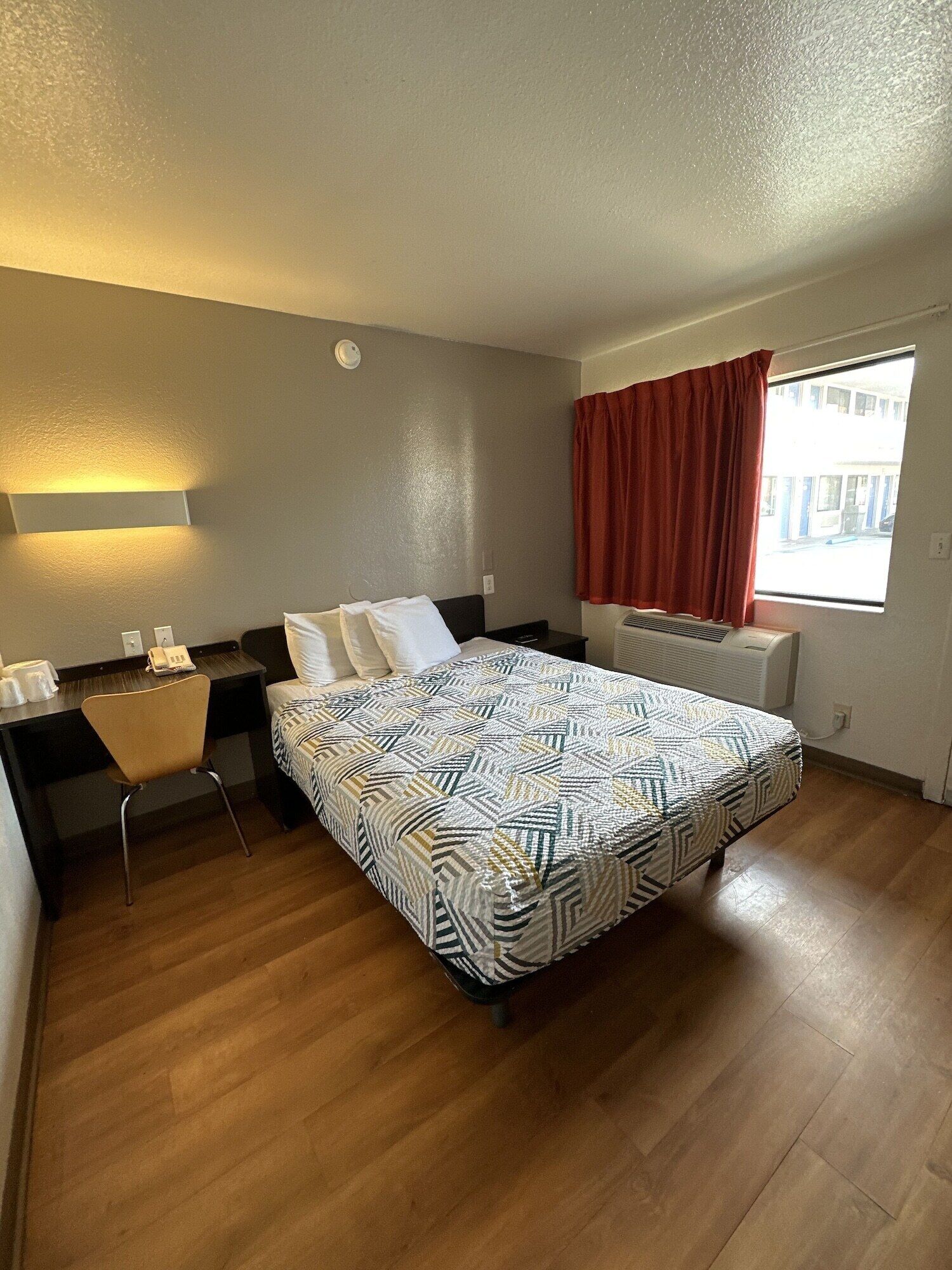 A hotel room with a bed, a desk, a chair, a window with curtains, and a radiator.
