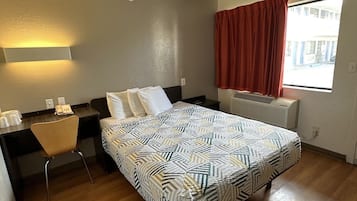 A hotel room with a bed, a desk, a chair, a window with curtains, and a radiator.