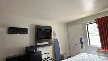 A hotel room with a bed, a desk with a television, a small ironing board, and a window with curtains.