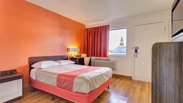 Ein Hotelzimmer mit Bett, Nachttisch, Lampe, Fenster mit Vorhängen und Klimaanlage.