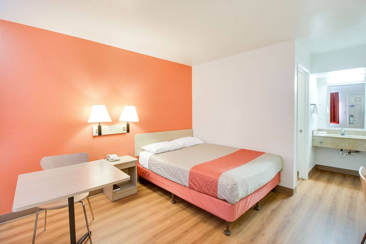 A hotel room with a bed, a desk, a chair, a nightstand, and a bathroom with a sink and mirror.