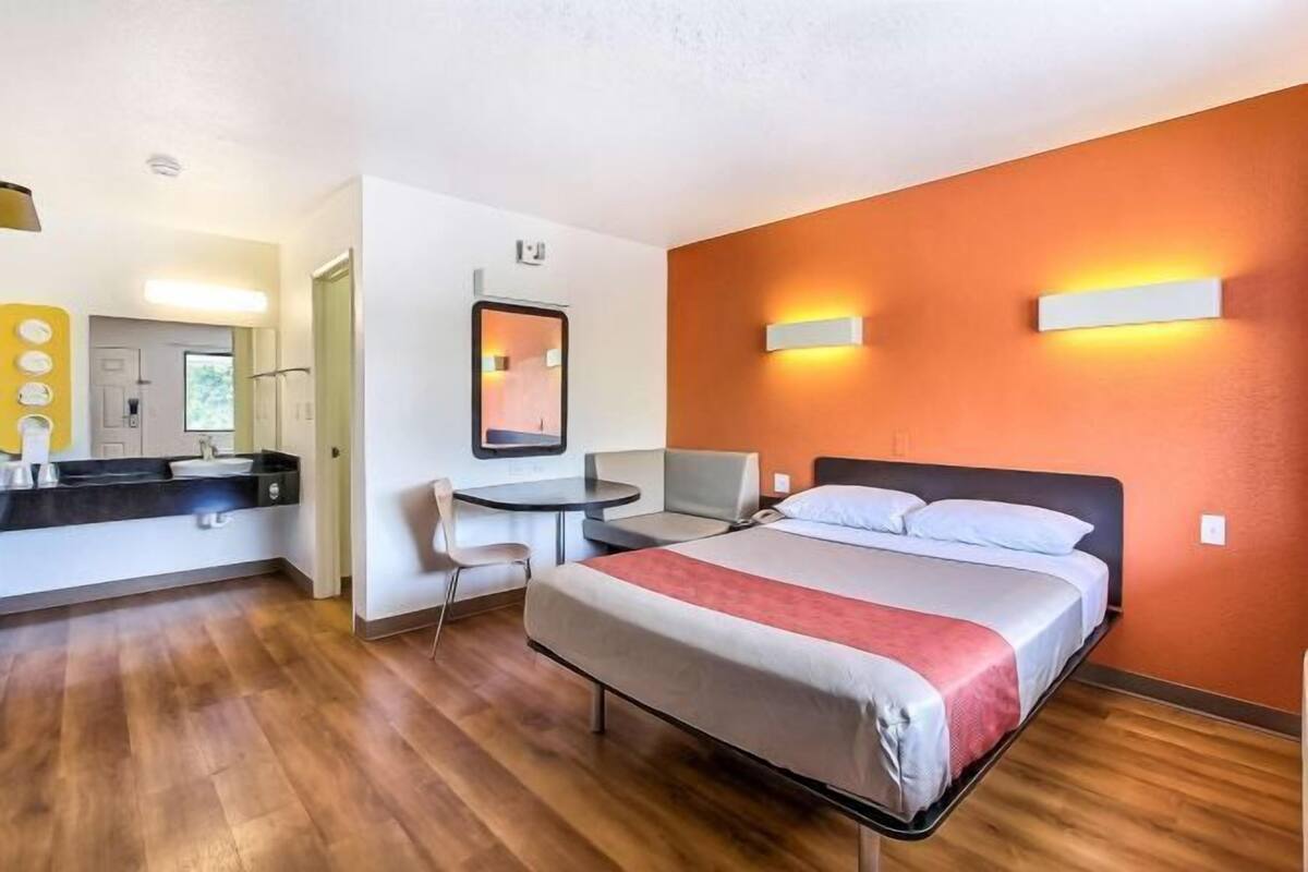 A hotel room with a bed, a desk, a chair, a mirror, and a bathroom with a sink and toilet.