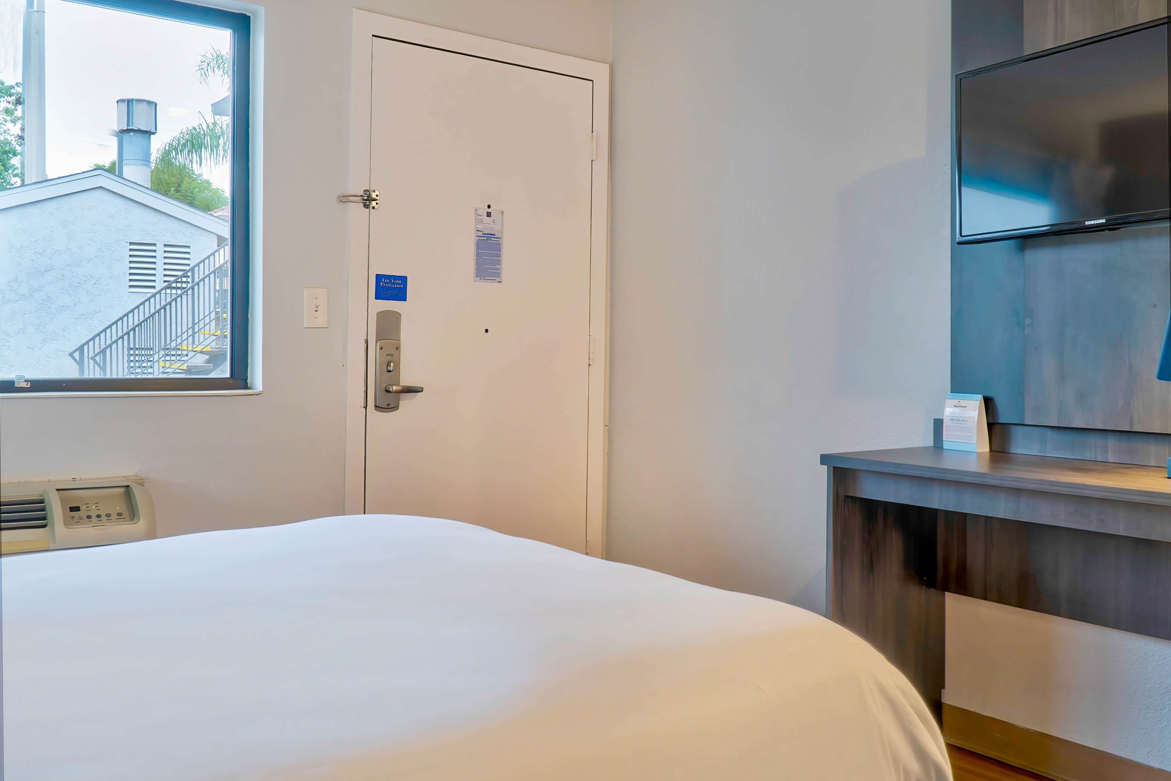 A hotel room with a bed, a door with a lock, a window with a view of a building, a wall-mounted TV, and a small table with a box on it.