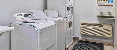 Un área de lavandería con lavadoras y secadoras apiladas, un panel de control, dispensador de blanqueador y suavizante, y una unidad de aire acondicionado con ventilación.