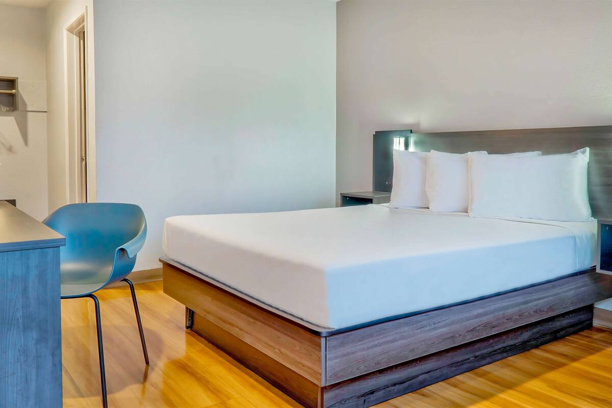 A modern hotel room with a wooden floor, a bed with white bedding, a blue chair, and a desk with a lamp.