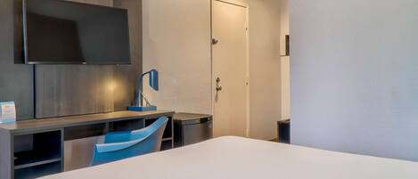 A hotel room with a bed, a desk with a chair, a TV mounted on the wall, and a door.