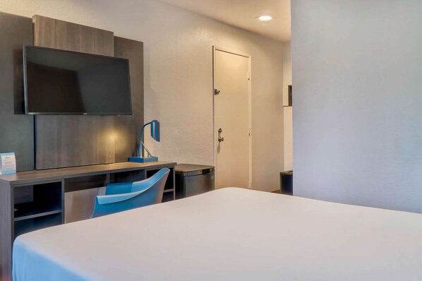 Una habitación de hotel con una cama, un escritorio con silla, un televisor montado en la pared y una puerta.