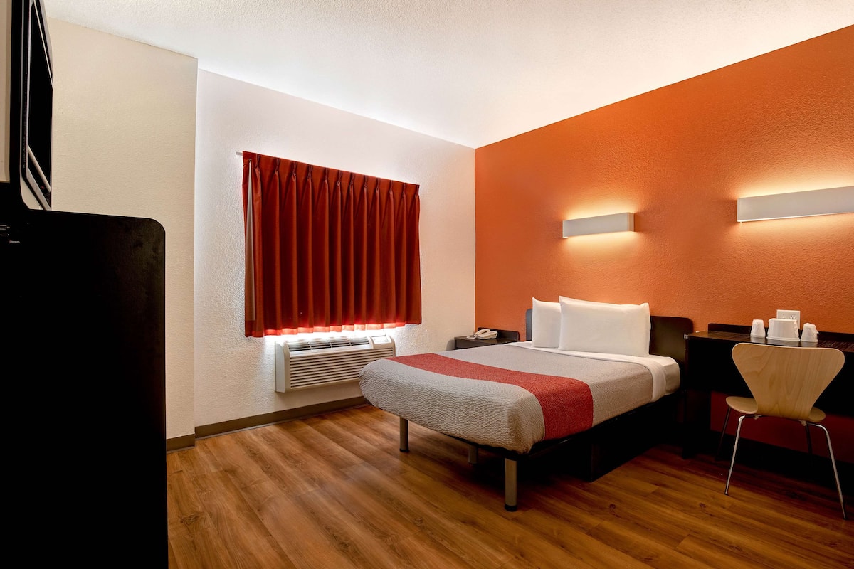Habitación de hotel con cama, escritorio, silla, ventana con cortinas y un calentador fijado a la pared.
