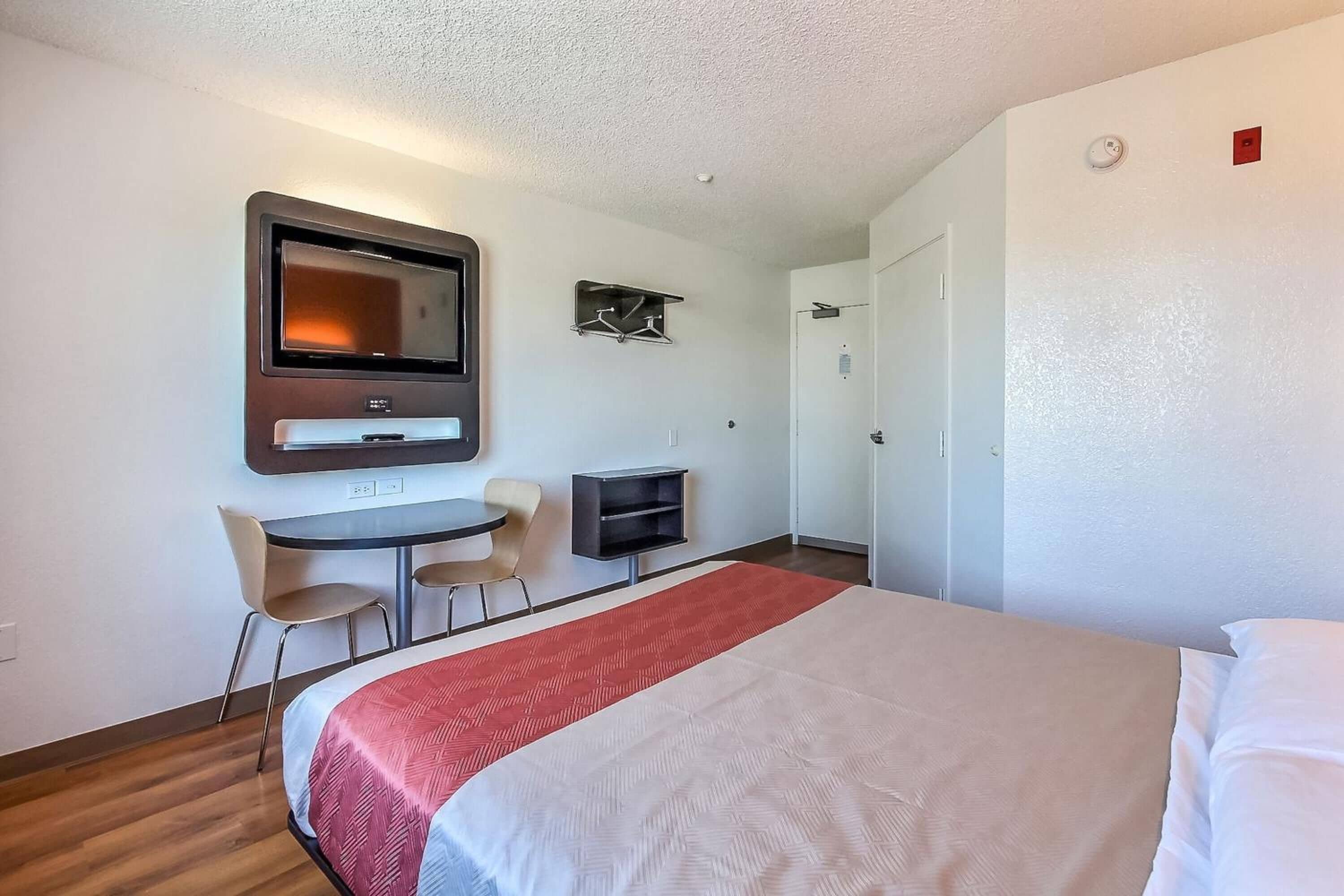 Quarto de hotel com cama, escrivaninha, cadeira, televisão e cabideiro.
