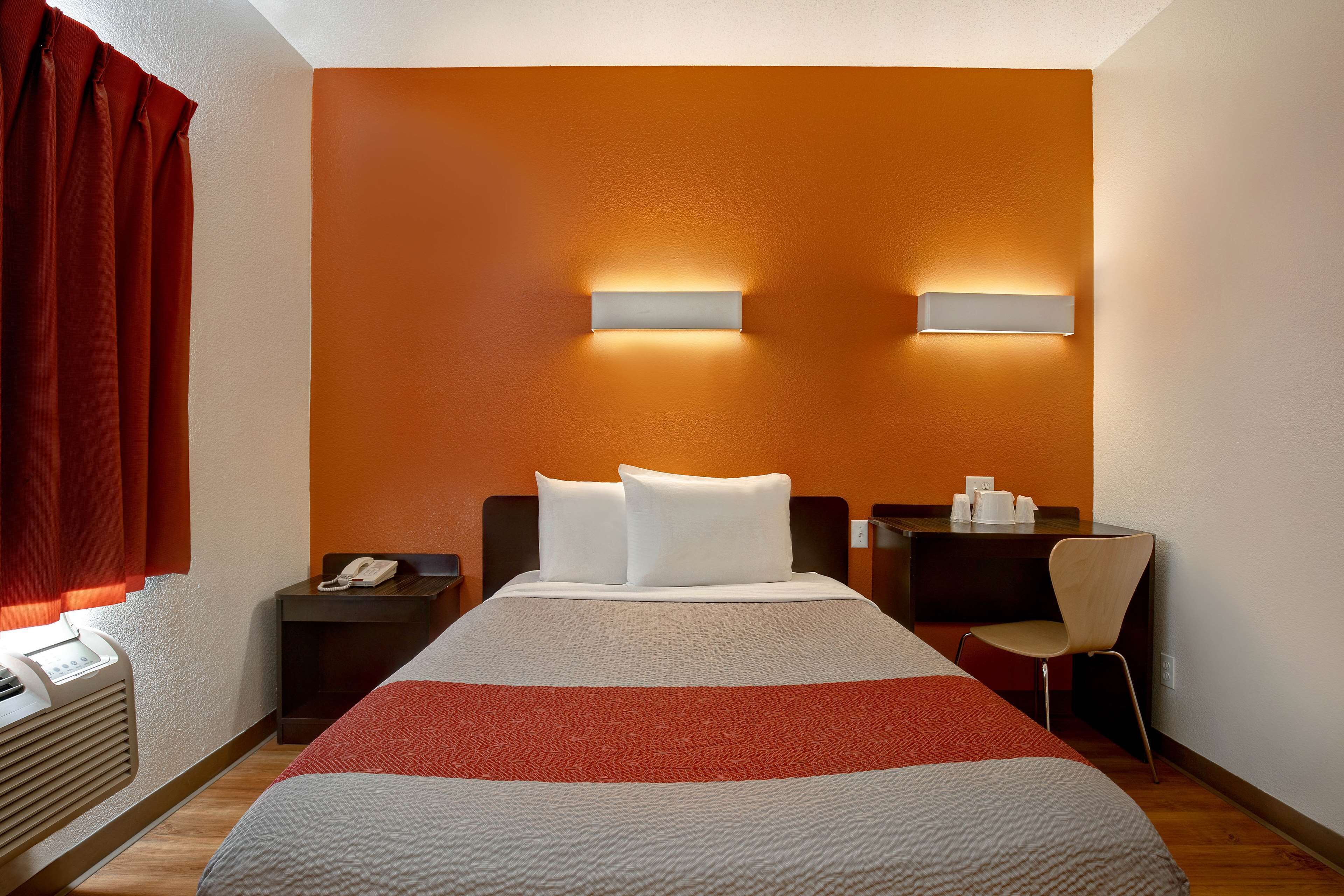 A hotel room with a bed, a desk, and a chair. There is a window with curtains, a wall-mounted phone, and a wall-mounted light fixture.