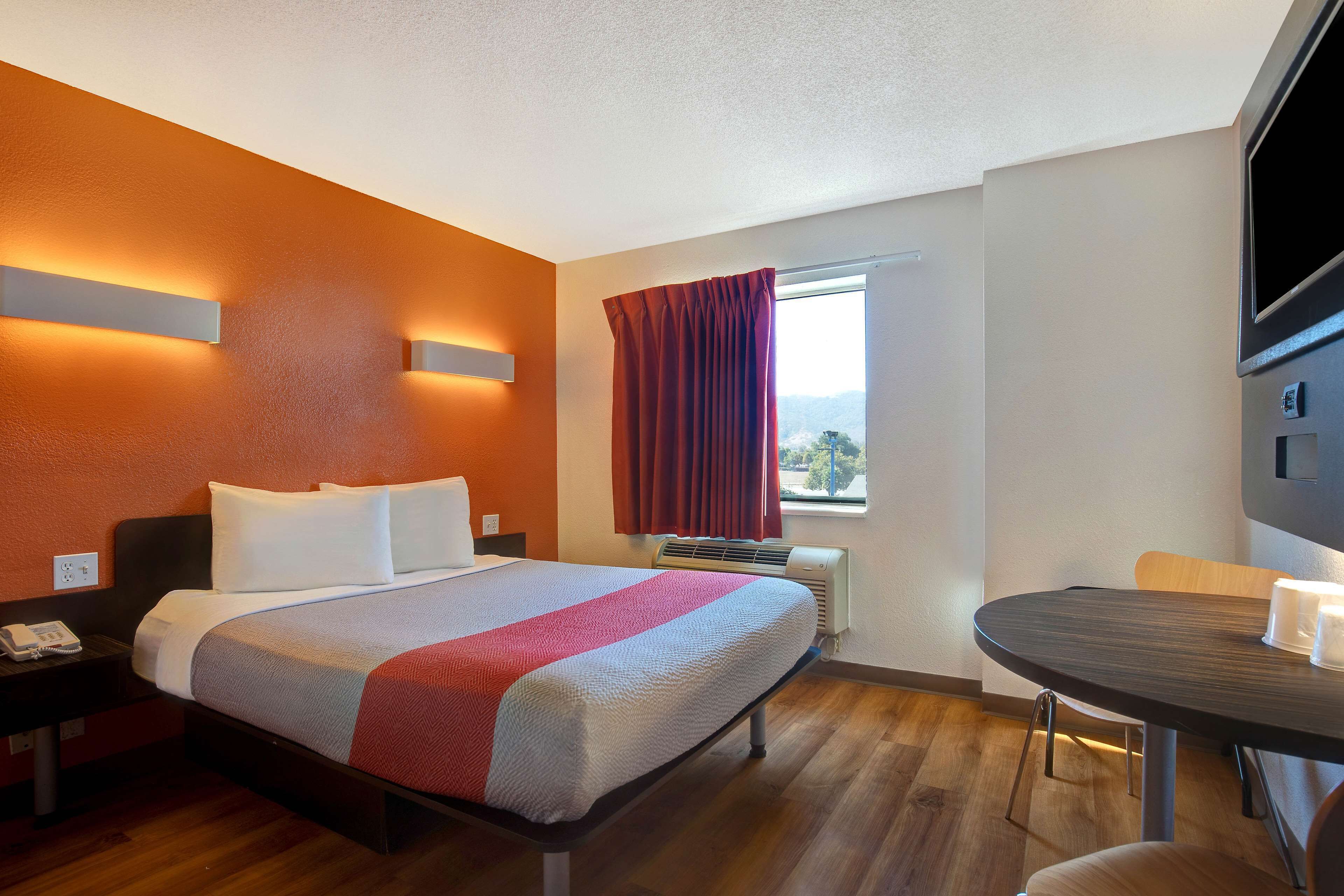 Habitación de hotel con cama, escritorio, silla, televisor y ventana con cortinas.