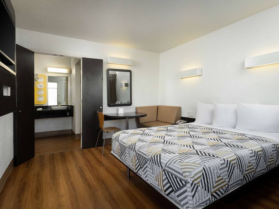 Une chambre d’hôtel avec un lit, un bureau, une chaise, une petite table, un miroir et une salle de bain visible à travers une porte ouverte.