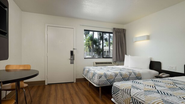 Ein Hotelzimmer mit zwei Betten, einem Schreibtisch, einem Stuhl und einem Fenster mit Blick auf Palmen.