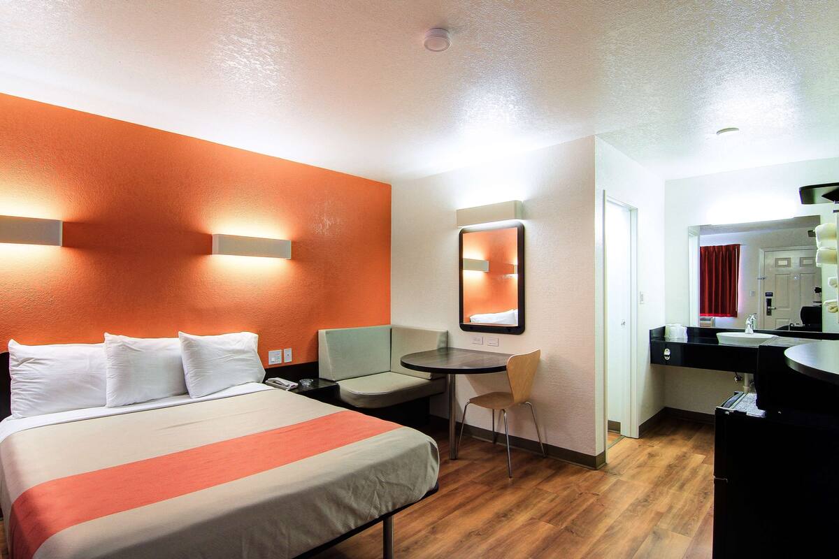 A hotel room with a bed, a desk, a chair, a small table, a mirror, and a bathroom visible through an open door.
