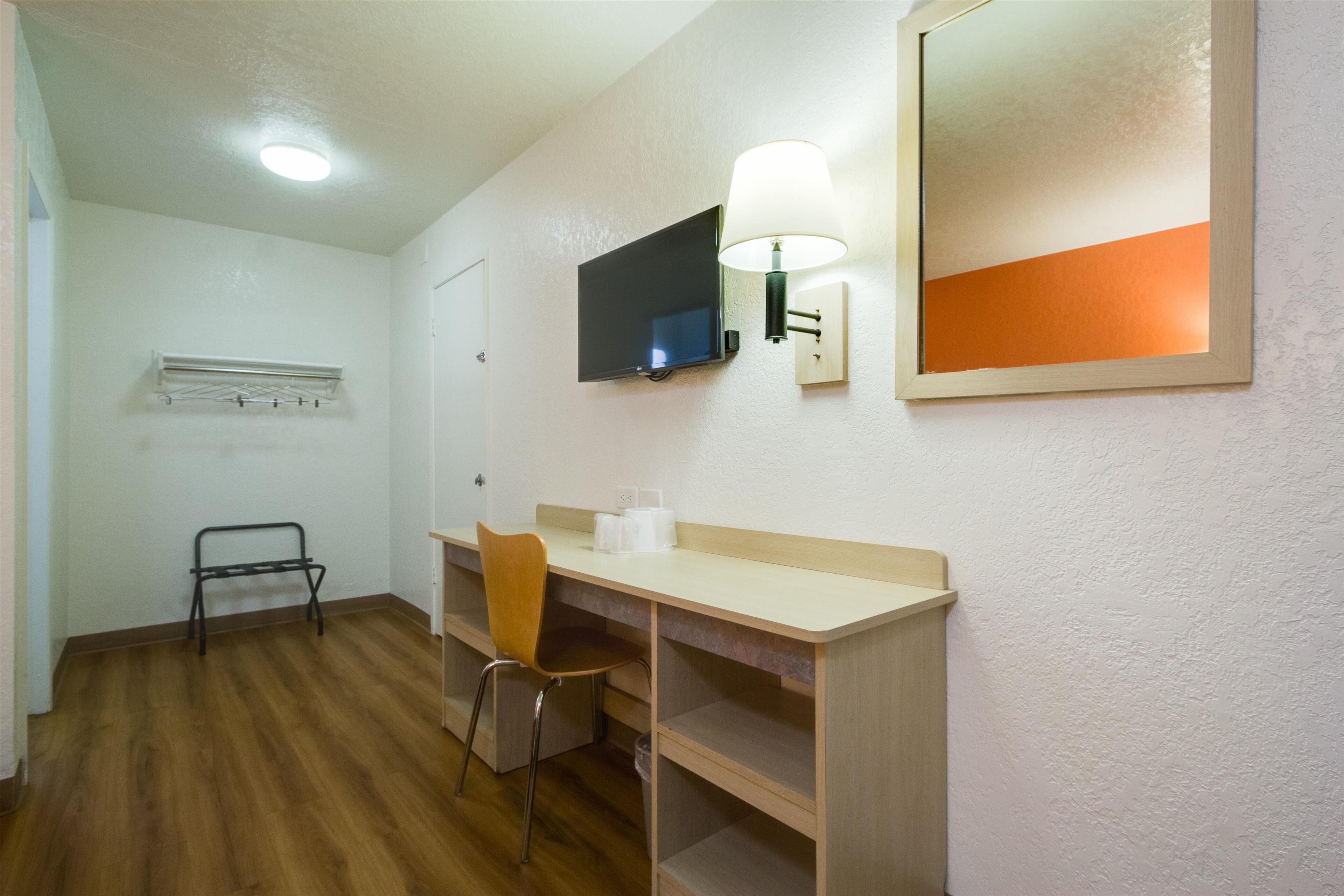 A small, well-lit room with a wooden desk, a chair, a television mounted on the wall, and a mirror above the desk.