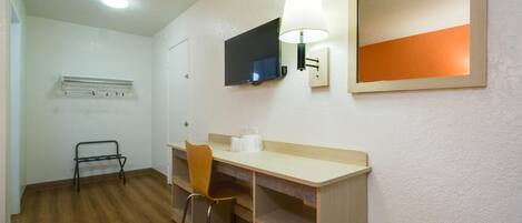 A small, well-lit room with a wooden desk, a chair, a television mounted on the wall, and a mirror above the desk.