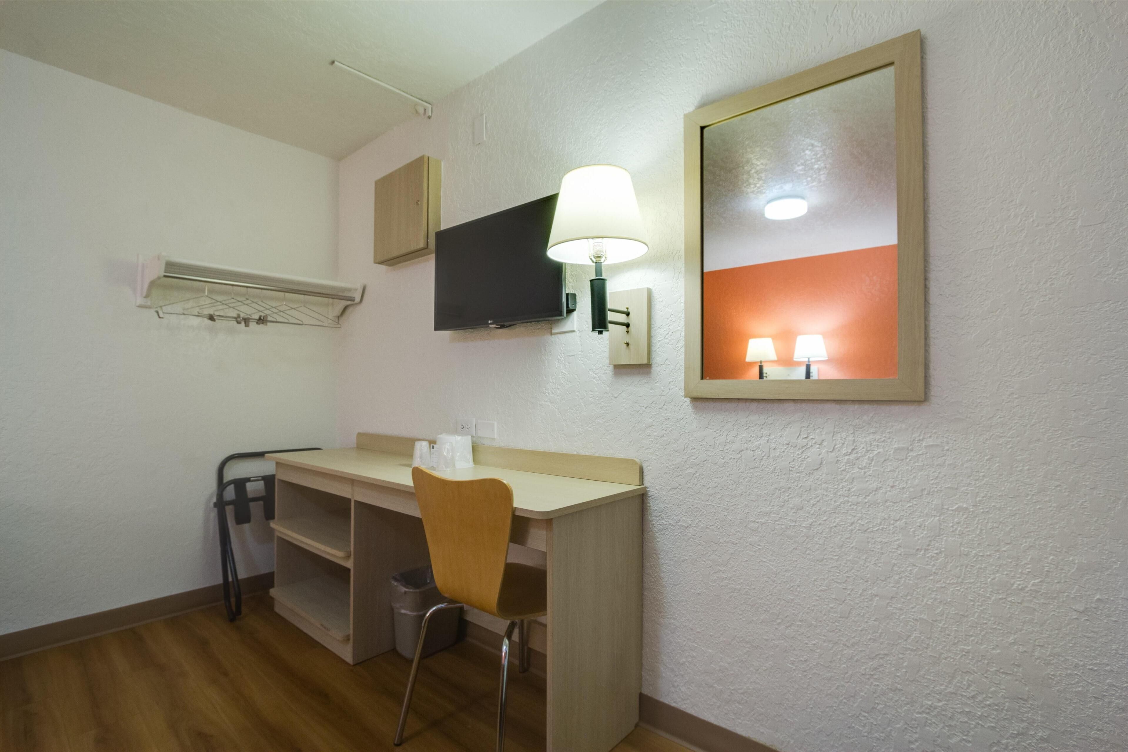 A small, well-lit room with a desk, chair, and a television mounted on the wall. There is a mirror and a lamp on the desk, and a small trash bin is visible.