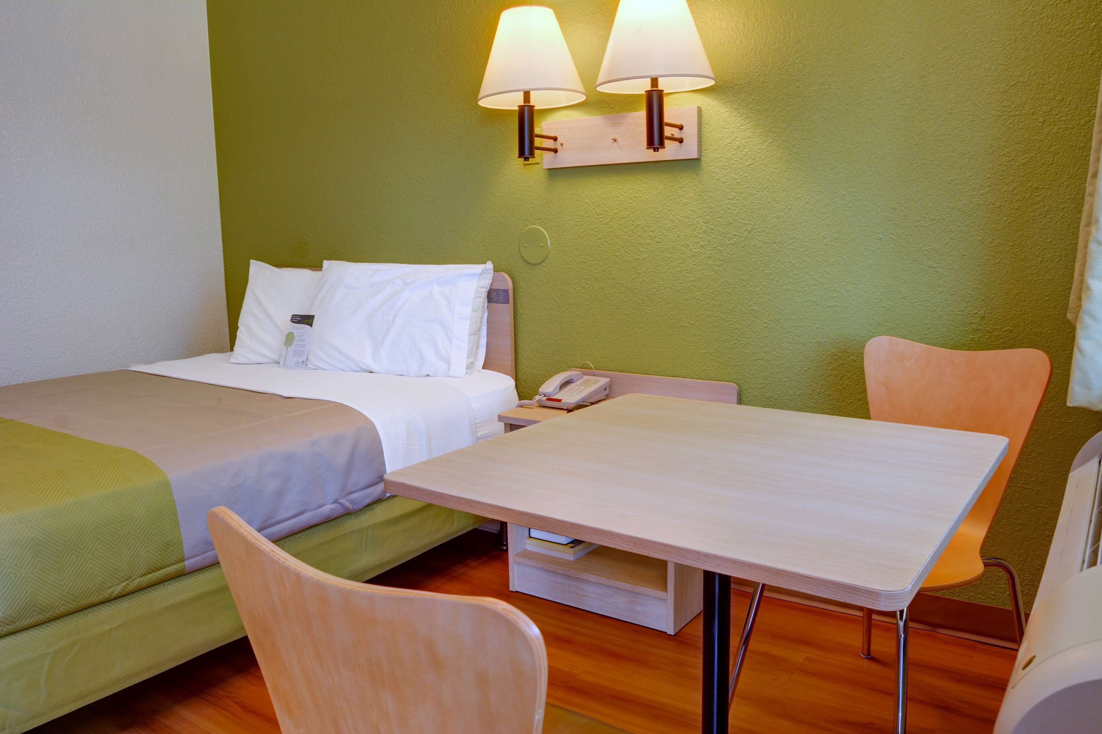 Hotellihuone, jossa on kaksi sänkyä, puinen pöytä, tuoli ja kaksi seinään kiinnitettyä lamppua.