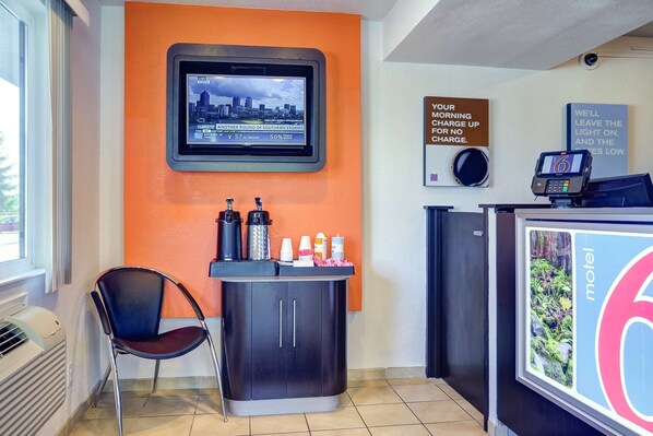 A hotel reception area with a TV displaying a cityscape, a counter with toiletries, a chair, and a sign about morning charge. - Motel 6 Eugene, OR - South Springfield (Eugene)