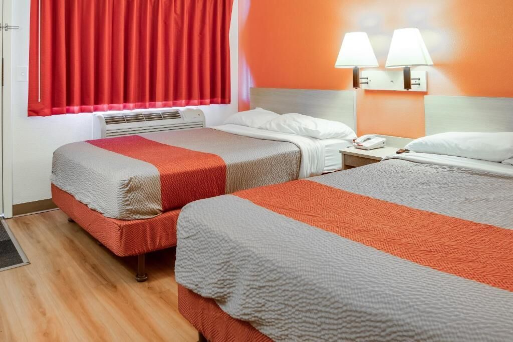 A hotel room with two beds, a heater, red curtains, and two wall lamps.