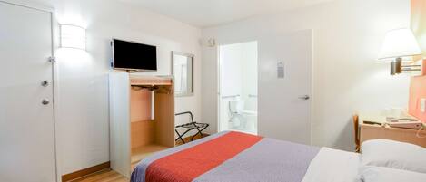 A hotel room with a bed, a TV mounted on the wall, a desk with a lamp, and a bathroom door.