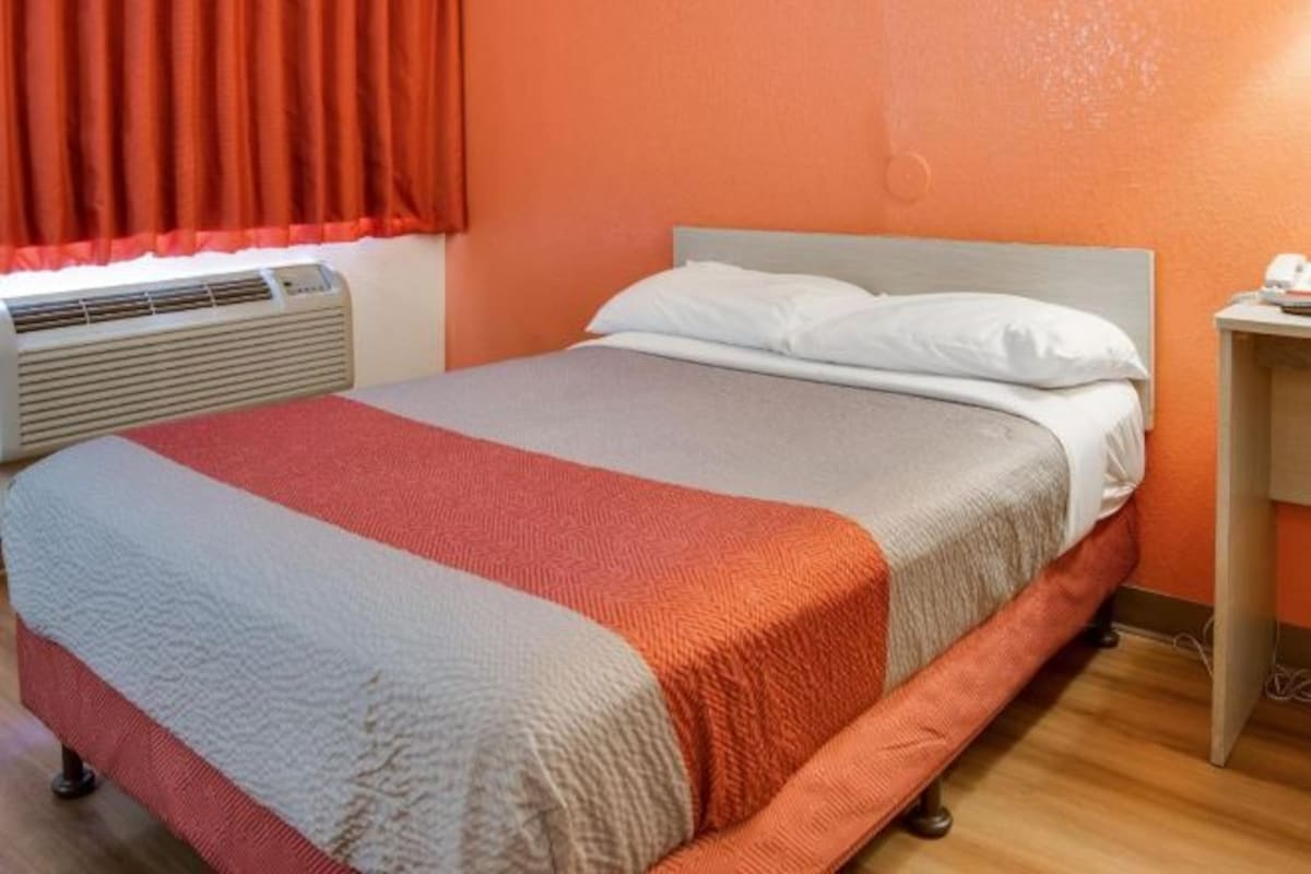 A single bed with a grey and red blanket, a white pillow, and a wooden nightstand with a lamp.