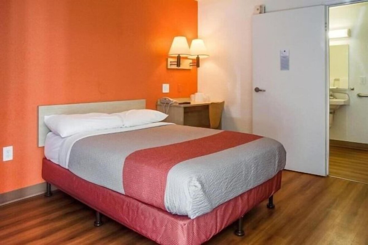 Un lit simple avec une couvre-lit rouge et blanche, une table de chevet avec une lampe et une porte donnant sur une salle de bain.