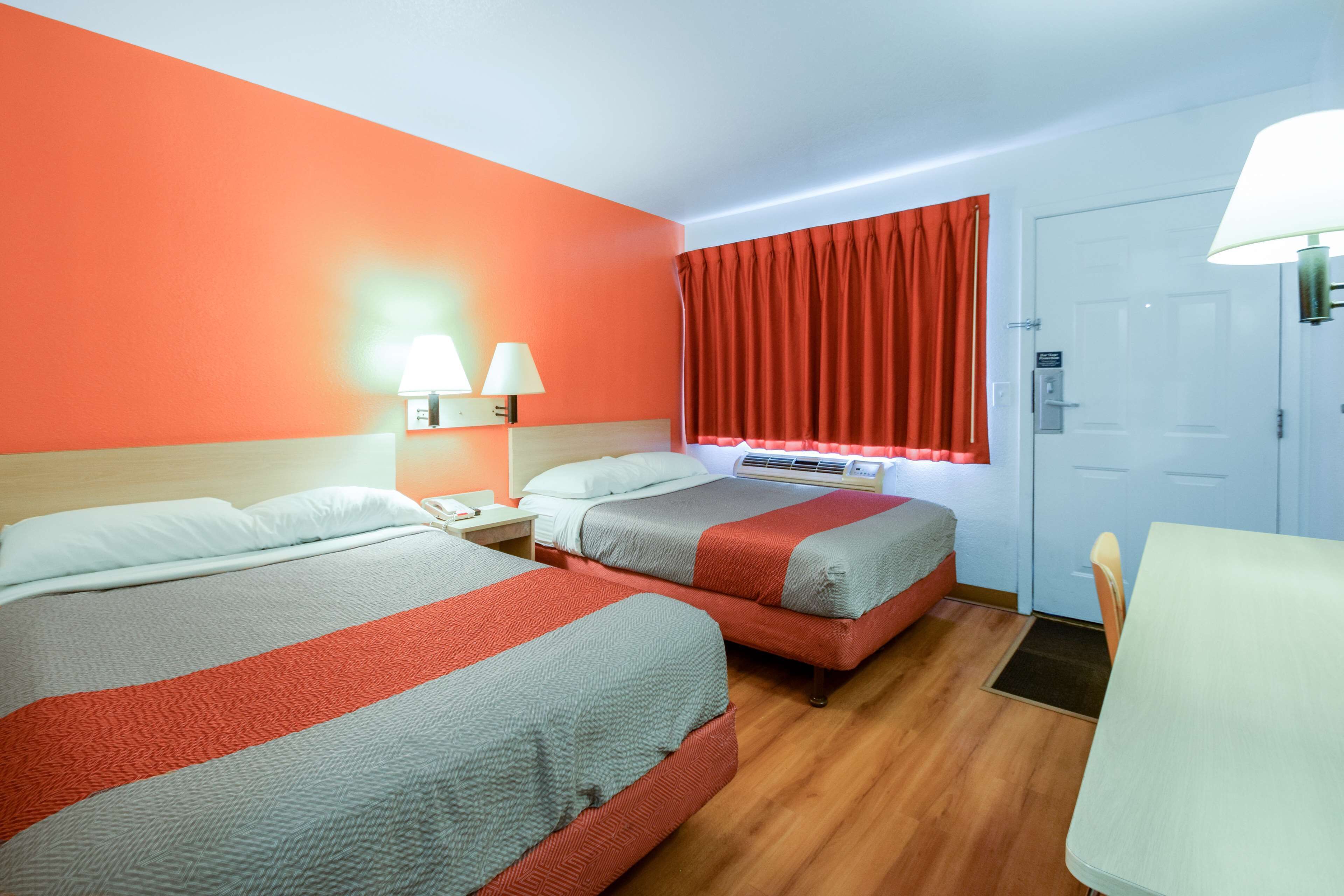 A hotel room with two beds, a desk, a chair, a window with red curtains, and a door.