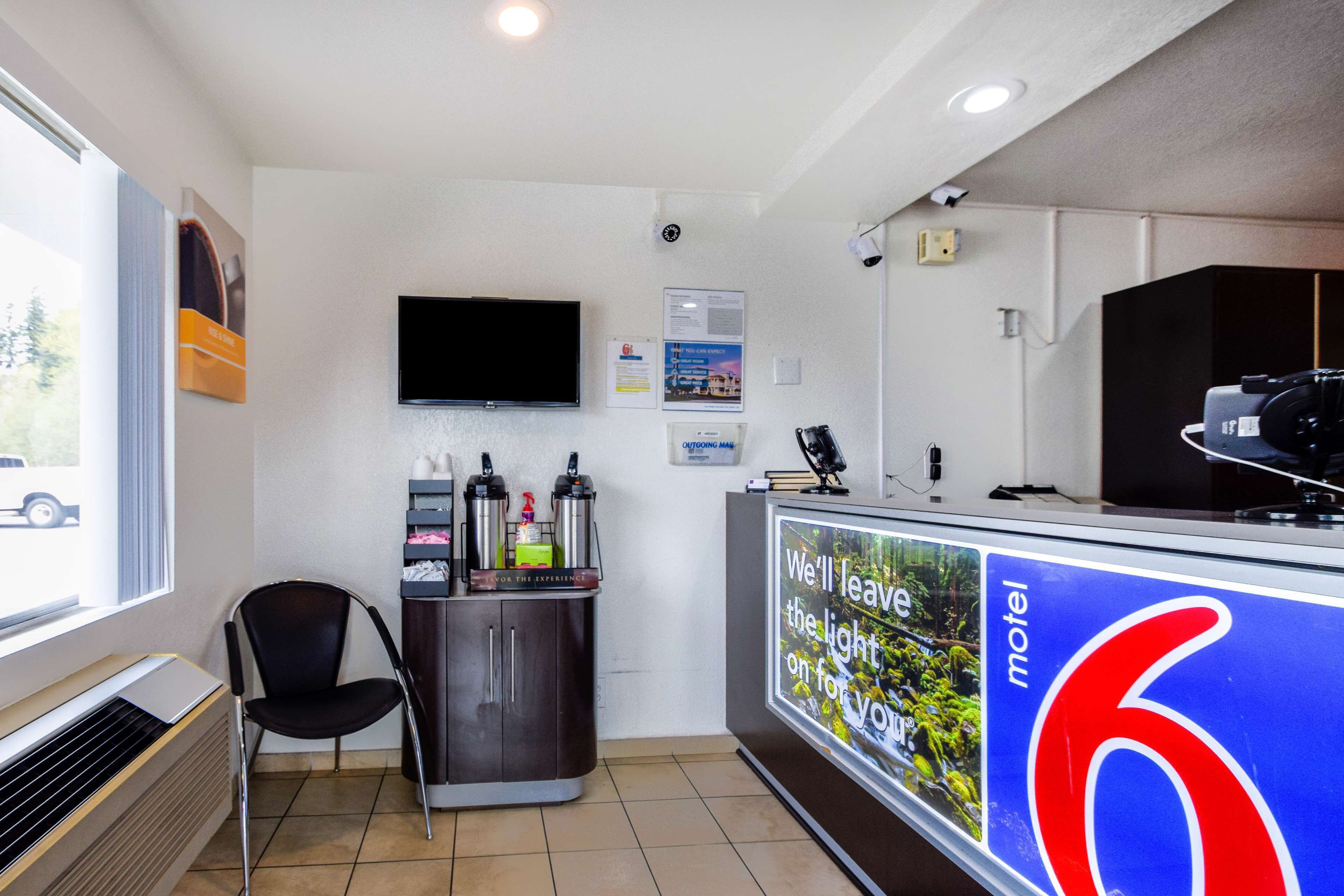 A motel reception area with a TV mounted on the wall, a counter with a large number '6' sign, a chair, and a vending machine.
