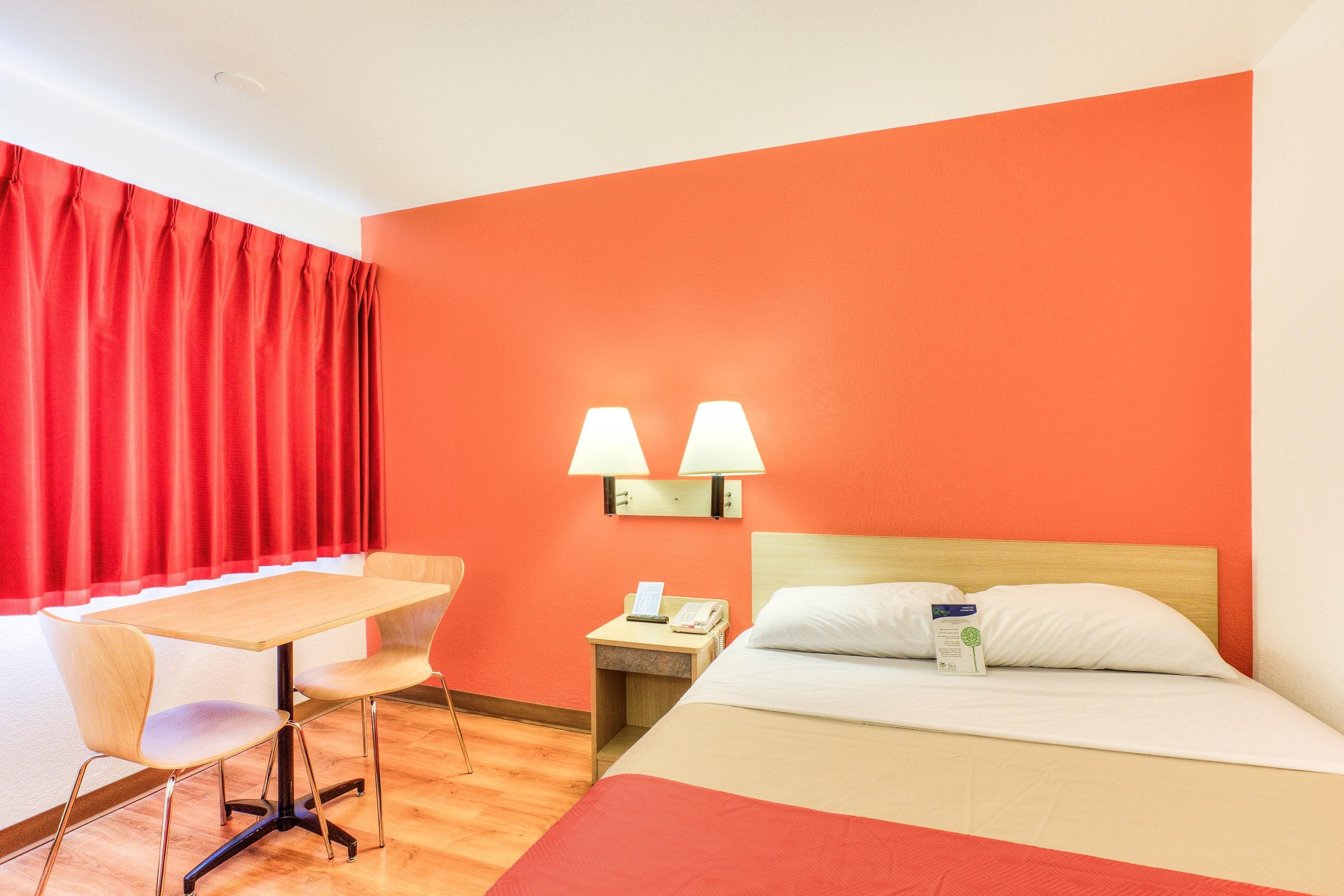 A hotel room with a wooden desk, two chairs, a bed with white bedding, a nightstand with a lamp, and a red curtain.