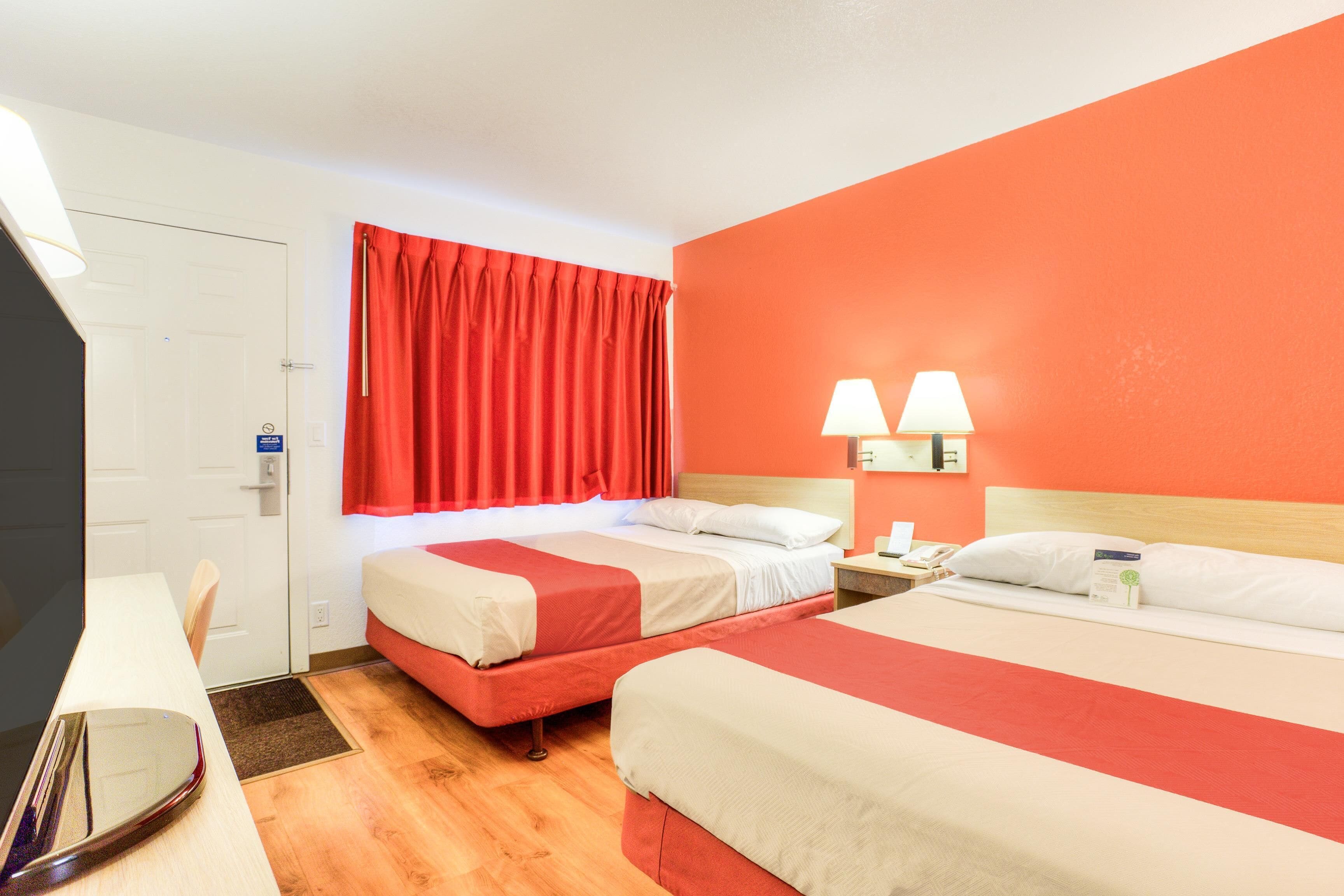 Quarto de hotel com duas camas, uma escrivaninha, um espelho e uma cortina vermelha.
