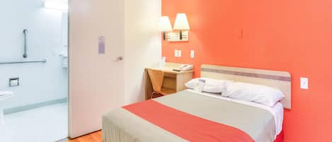 Ein Hotelzimmer mit Bett, Schreibtisch, Stuhl, Nachttisch, Lampe und einem Badezimmer mit Dusche.
