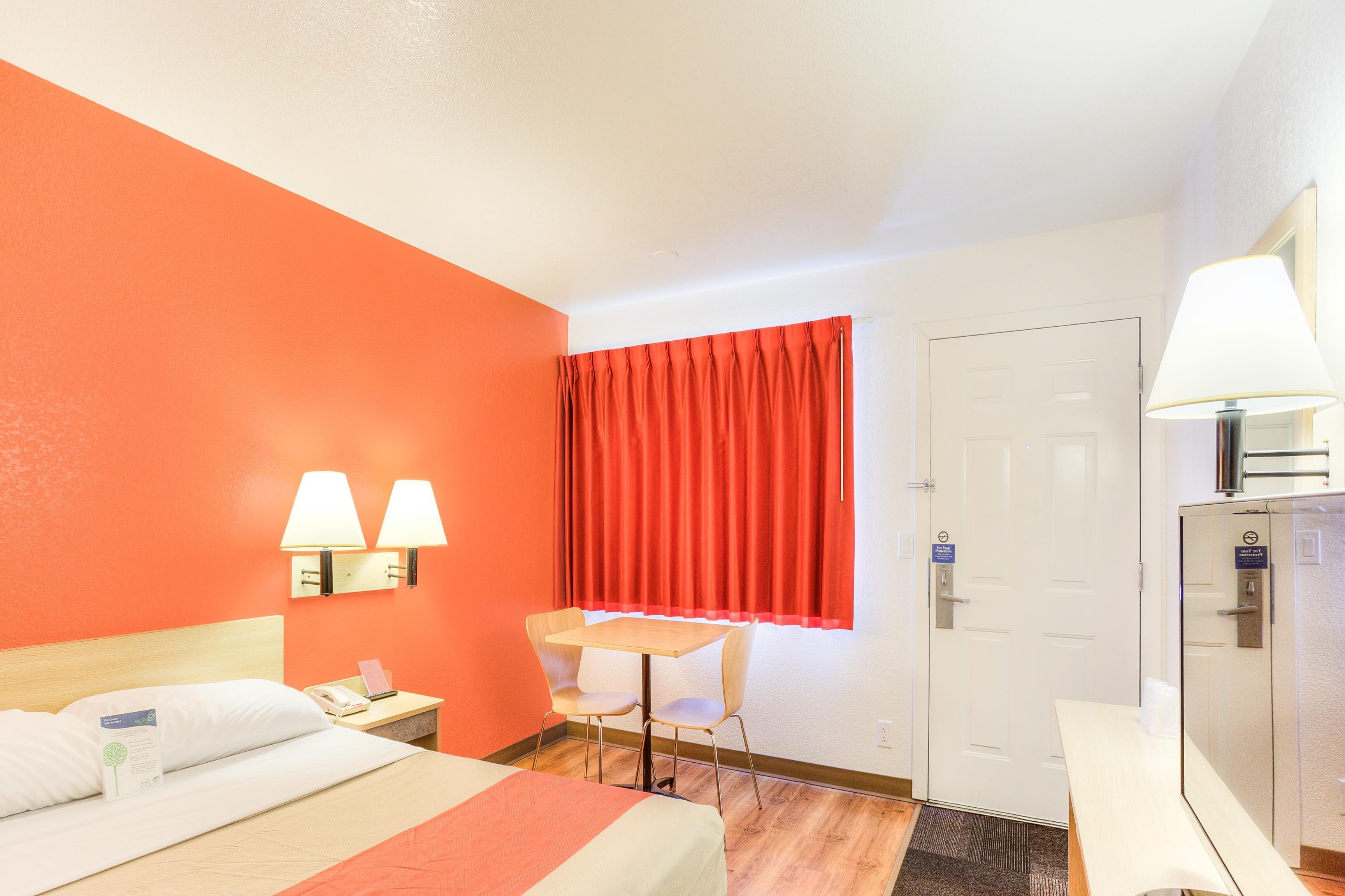 A hotel room with a bed, a desk, a chair, a window with red curtains, and a door.